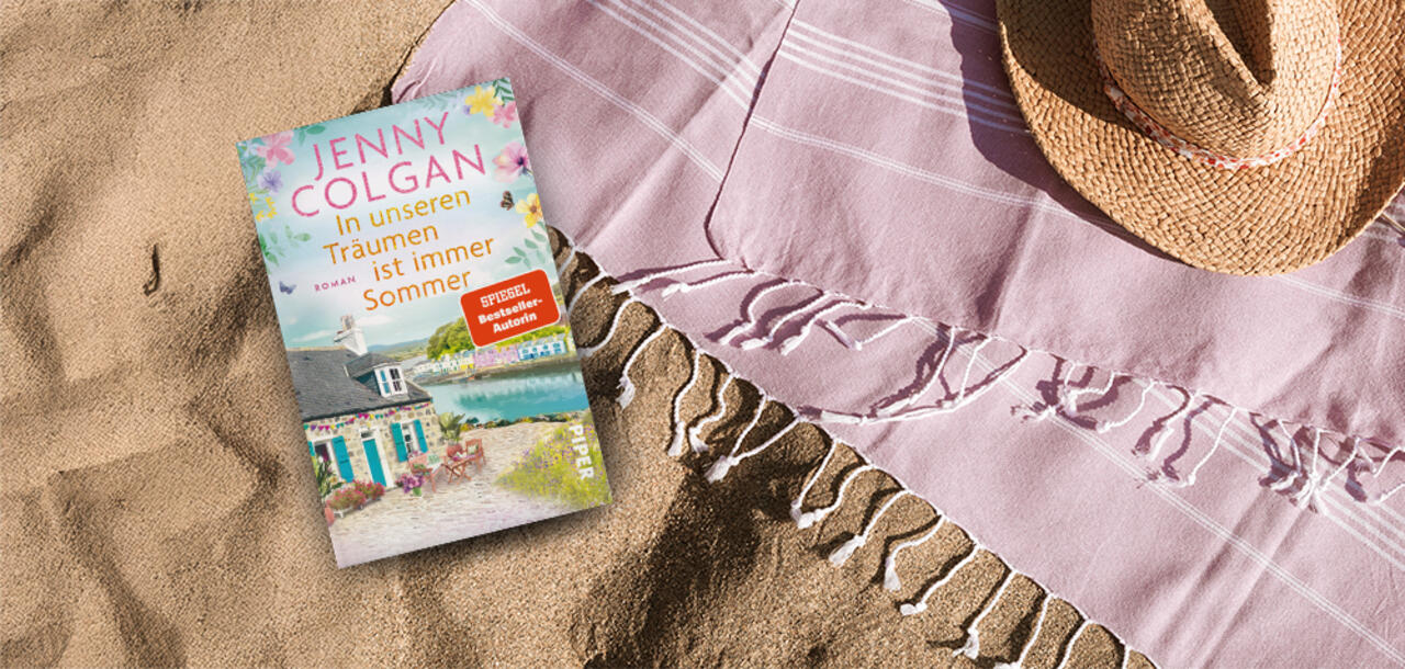 Buch  'In unseren Träumen ist immer Sommer' auf Sand neben pinkem Tuch und Strohhut.