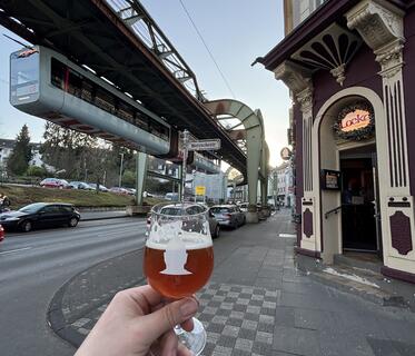 Ein Stielglas mit bernsteinfarbenem Bier wird von einer Hand vor die Wuppertaler Schwebebahn gehalten. Im Hintergrund sind die Haltestelle „Nietzschestraße“, parkende Autos und der Eingang zur Kneipe „Locke“ sichtbar.