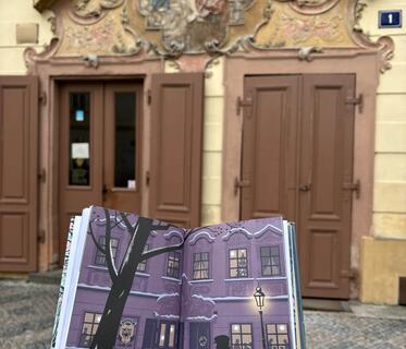 Ein Buch mit einer gezeichneten Straßenszene wird von einer Hand vor ein verziertes Gebäude gehalten, das der Zeichnung ähnelt.