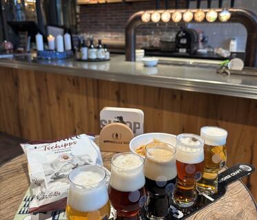 Eine Auswahl an fünf verschiedenen Bieren steht auf einem Verkostungstablett auf einem Holztisch. Daneben befindet sich eine weiße Chipstüte, ein Bierdeckelhalter und eine Karte mit der Aufschrift „Appenzeller Bier“. Im Hintergrund sind unscharf die Bar der Brauerei mit Zapfhähnen und ein Regal mit Flaschen zu sehen.