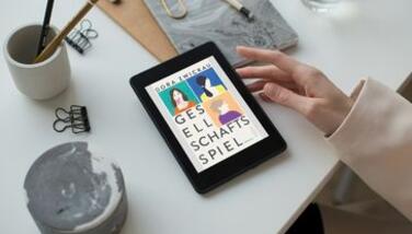 Eine Hand berührt ein Tablet, das auf einem weißen Tisch liegt. Der Bildschirm zeigt das Buchcover "Gesellschaftsspiel"
