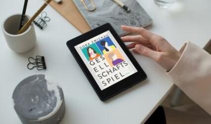 Eine Hand berührt ein Tablet, das auf einem weißen Tisch liegt. Der Bildschirm zeigt das Buchcover "Gesellschaftsspiel"