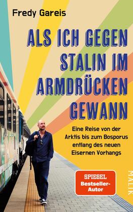 Das Buchcover zeigt einen bärtigen Mann in einem blauen Anzug, der an einer Bahnstation steht. Er blickt nachdenklich in die Ferne. Im Hintergrund verläuft ein Zug, und der Himmel ist in sanften Farben mit gelb, orange, grün und blau abgebildet, die eine dynamische Atmosphäre schaffen. Der Titel „ALS ICH GEGEN STALIN IM ARMDRÜCKEN GEWANN“ steht in großen, fetten weißen Buchstaben, während der Untertitel „Eine Reise von der Arktis bis zum Bosporus entlang des neuen Eisernen Vorhangs“ in etwas kleinerer Schrift darunter platziert ist.