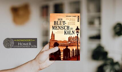 "Der Blutmensch zu Köln" wird von einer Hand hochgehalten. Im Hintergrund ist unscharf ein Wohnraum mit einem Bücherregal zu sehen. Links im Bild ist der Nominiert-Button für den Homer 2026 zu sehen.