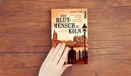 „Der Blutmensch zu Kökn“ liegt auf einem Holztisch und wird von einer Hand gehalten.