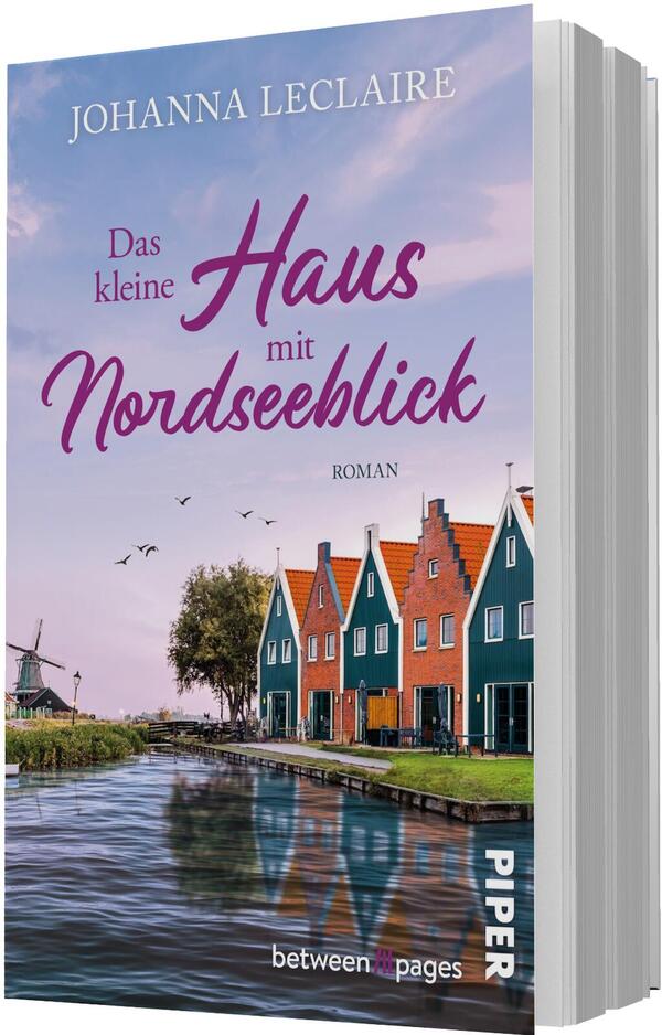 Das Buchcover zeigt eine ruhige Nordseekulisse mit bunten, schmalen Giebelhäusern (türkis, orange, rot) an einem Kanal, deren Spiegelungen im Wasser sichtbar sind, eine Windmühle links und ziehende Vögel vor einem pastellrosa bis violettem Abendhimmel. Oben steht der Autorenname Johanna Leclaire, der Titel „Das kleine Haus mit Nordseeblick“ ist in großer, verspielter magenta Script‑Typografie, darunter klein „ROMAN“.