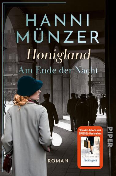 Das Buchcover zeigt eine melancholische Straßenszene in Schwarz-Weiß, die von historischen Gebäuden umrahmt ist. Im Vordergrund steht eine Frau mit blauer Mütze und grauem Mantel, die den Rücken zur Kamera gewandt hat. Graue Schatten von Passanten ziehen sich über den Boden. Der Titel „Honigland“ und der Untertitel „Am Ende der Nacht“ sind in eleganter, hellblauer Typografie platziert, während der Name der Autorin Hanni Münzer darüber hervorgehoben ist. Unten links befindet sich ein Hinweis auf die Bestseller-Autorin.