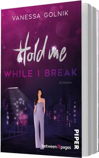 Das Buchcover zeigt einen nächtlichen, violett-pinken Stadtblick mit glitzernden Lichtern. Oben steht der Autorenname Vanessa Golnik in Großbuchstaben. Der Titel „Hold me“ ist groß in silbrig-weißem, handschriftlichem Schriftzug, darunter in kräftigem Violett „WHILE I BREAK“ in Versalien. Vor der Skyline steht eine elegante, dunkelhaarige Frau in lila Jumpsuit, frontal, die Stimmung ist melancholisch und zugleich glamourös.