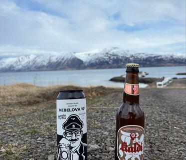 Eine Bierdose mit dem gezeichneten Gesicht eines Mannes, der eine Bahnmütze trägt und einen Bierkrug hält, sowie eine Flasche mit isländischem „Kaldi“-Bier stehen auf einem Kiesweg. Im Hintergrund sind ein See und schneebedeckte Berge zu sehen.