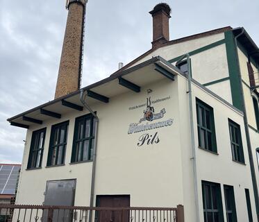 Die Wittichenauer Brauerei zeigt eine helle Fassade mit dunkelgrünen Fenstern und dem Schriftzug der Brauerei. Man sieht links einen hohen runden Schornstein auf dem Dach und in der Mitte einen eckigen, auf dem sich ein Storchennest befindet.