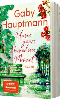 Das Buchcover zeigt eine idyllische Gartenszene mit bunten Blumen in Rottönen und grünem Laub. In der oberen Hälfte prominent platziert, steht der Name der Autorin „Gaby Hauptmann“ in einer lebhaften Schriftart. Darunter befindet sich der Titel „Unser ganz besonderer Moment“ in geschwungener, roter Schrift. Am unteren Rand ist das Wort „Roman“ in einer schlichten Schriftgröße zu sehen. Die Farbtöne vermitteln eine harmonische und einladende Atmosphäre.