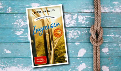 „Zugreisen“ von Jaroslav Rudiš liegt auf einer hellblauen, verwitterten Holzfläche mit einem Seilknoten daneben.