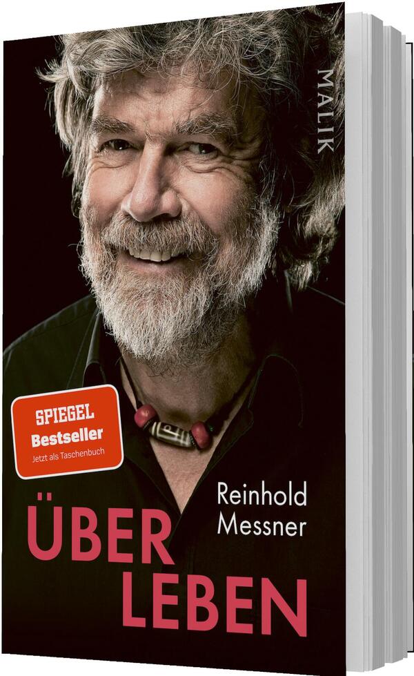 Das Buchcover zeigt ein großformatiges Porträt des lächelnden, freundlichen Reinhold Messner vor schwarzem Hintergrund; graues, zerzaustes Haar und Vollbart, sichtbare Hautfalten, braunes Hemd, Halskette mit roten Perlen. Titel „ÜBERLEBEN“ in kräftigem Pink, groß, versetzt über zwei Zeilen; Autorenname in weißer, schlichter Schrift. Auf der Vorderseite ein roter Button mit der Aufschrift „SPIEGEL Bestseller“.