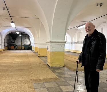 Ein weißhaariger Mann mit Gehstock steht in einem historischen Brauereiraum mit Deckengewölbe. Auf dem Boden liegt Getreide, das in flachen, quadratischen Haufen sortiert wurde.