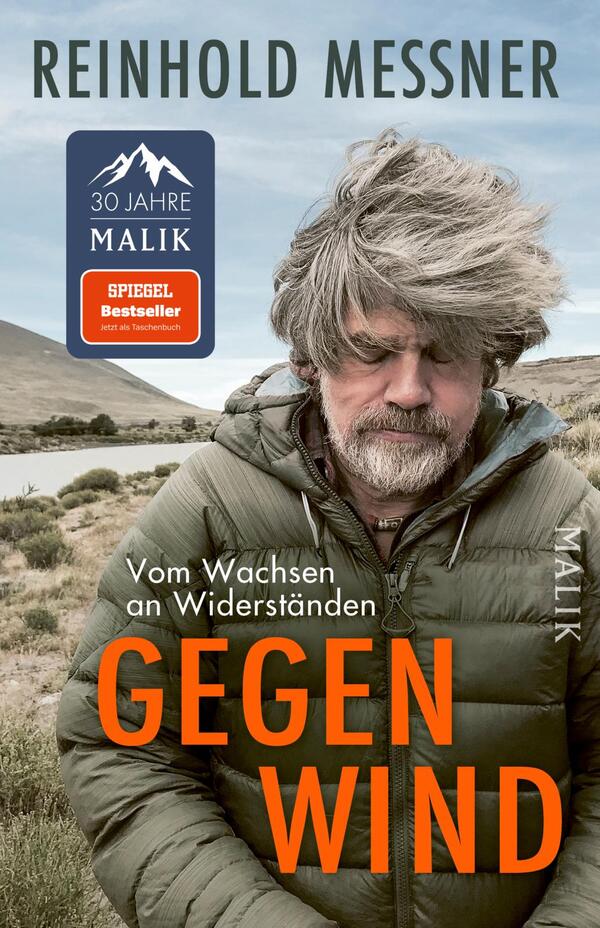 Das Buchcover zeigt ein Porträt von Reinhold Messner, der in einem gebräuntem Parka vor einer kargen Landschaft mit sanften Hügeln und einem Gewässer steht. Sein zerzaustes, graues Haar vermittelt eine Atmosphäre der Ungezwungenheit und Stärke. Der Titel „GEGEN WIND“ ist in auffälliger, orangefarbener Schrift in der unteren Mitte platziert, während der Untertitel „Vom Wachsen an Widerständen“ und der Verlag „MALIK“ in schlichterer Schrift darüber stehen.