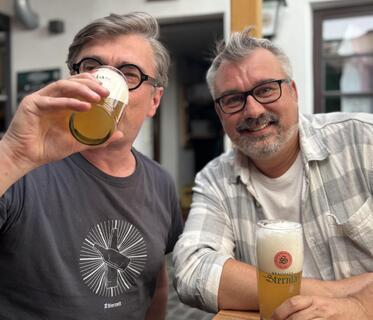 Der Autor und eine weitere Person sitzen an einem Holztisch, der im Hof vor einem Gebäude mit geöffneter Tür steht. Der Autor trinkt helles Bier aus einem Glas mit der Aufschrift „Brauhaus Sternla“. Vor dem zweiten Mann steht so ein Glas auf dem Tisch.