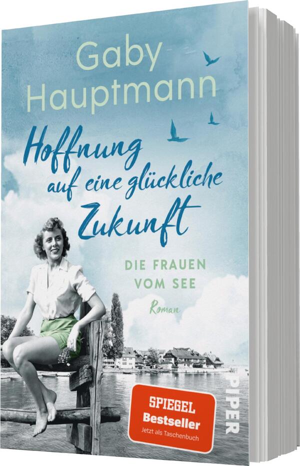 Das Buchcover zeigt ein hellblaues, wolkiges Himmelshintergrund mit einem kleinen See und grünen Ufern, auf dem eine Frau in heller Bekleidung sitzt. Sie blickt optimistisch in die Ferne. Der Titel „Hoffnung auf eine glückliche Zukunft“ steht in geschwungener Schrift in Blau und ist prominent platziert. Darunter steht in leichterer Schrift „Die Frauen vom See“. Der Autorinnenname „Gaby Hauptmann“ ist ebenfalls in blauer Schrift oberhalb des Titels platziert. Ein rotes Banner mit dem Text „SPIEGEL Bestseller“ befindet sich unten rechts.