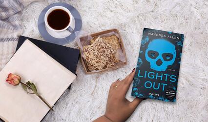 Auf einer flauschigen Unterlage liegt das Buch „Lights Out“, daneben eine Tasse Tee auf Untertasse, eine Schachtel mit Keksen und ein geöffnetes Buch, in dem eine getrocknete Rose liegt.