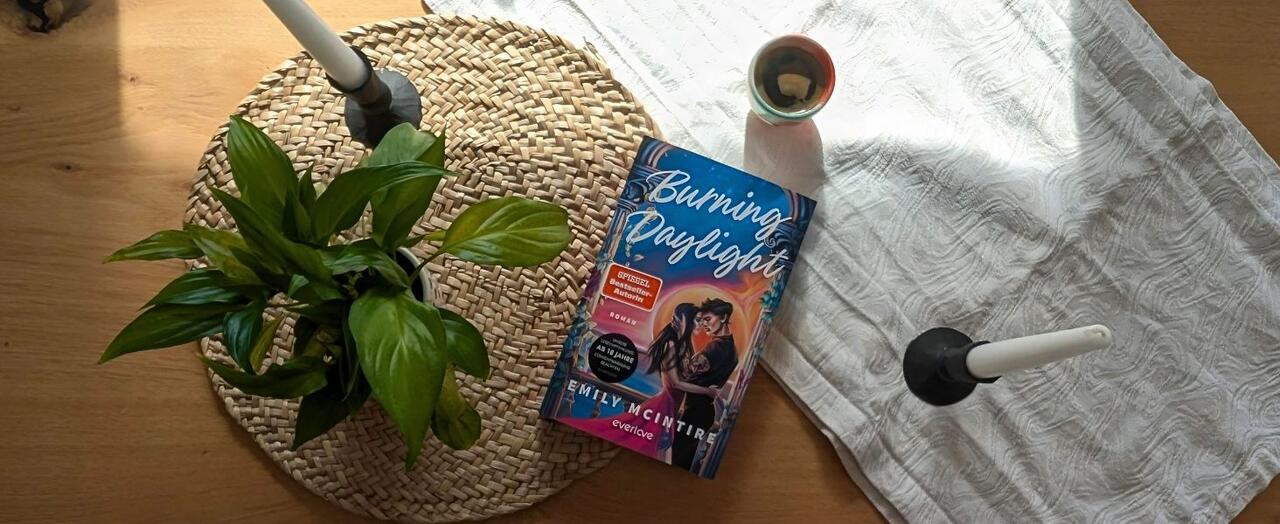 „Burning Daylight“ liegt auf einem Tisch. Darunter liegen ein Tischset aus Bast und ein weißer Tischläufer. Daneben stehen eine kleine Topfpflanze sowie zwei Stabkerzen.