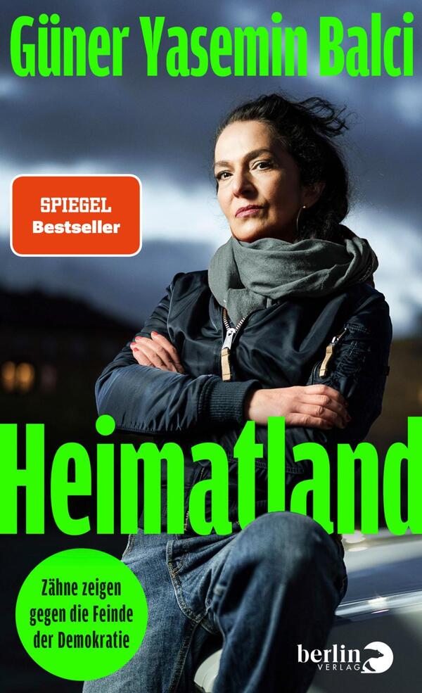 Das Buchcover zeigt eine Porträtaufnahme einer Frau, die ernst in die Kamera blickt. Sie trägt einen schwarzen Winterjacke und eine graue Schal. Der Hintergrund ist in düsteren, graublauen Tönen gehalten, was eine nachdenkliche Stimmung erzeugt. Der Titel „Heimatland“ ist in großen, leuchtend grünen Buchstaben platziert. Darunter steht der Untertitel „Zähne zeigen gegen die Feinde der Demokratie“ in einer runden, grünen Blase. Der Autorinnenname „Güner Yasemin Balci“ ist oben auf dem Cover in schlichtem Weiß abgedruckt.
