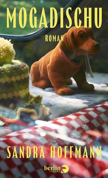 Das Buchcover zeigt ein sonnenbeleuchtetes Picknickmotiv: ein braunes Stoffdackelspielzeug sitzt auf einem gepolsterten Untergrund, vorne liegt ein rot-weiß kariertes Tuch, links ein gelb-grün gestrickter Hut. Im unscharfen Hintergrund grünes Laub. Der Titel „MOGADISCHU“ steht oben in großen gelben Großbuchstaben, darunter klein „ROMAN“, und unten die Autorin „SANDRA HOFFMANN“. Stimmung: warm und nostalgisch.