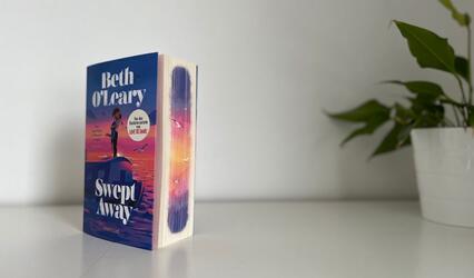 „Swept away“ von Beth O'Leary steht auf einem weißen Tisch vor einer weißen Wand. Am rechten Bildrand ist eine Topfpflanze zu sehen.