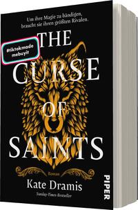 Das Buchcover zeigt einen schwarzen Hintergrund, auf dem ein majestätischer, golden gefärbter Wolfskopf in der Mitte abgebildet ist. Darüber prangt in großen, weißen Buchstaben der Titel „THE CURSE OF SAINTS“. Darunter steht in kleinerer Schrift „Roman“ und der Name der Autorin, Kate Dramis, gefolgt von der Angabe „Sunday-Times-Bestseller“. Ein bunter Sticker mit der Aufschrift „#tiktokmade“ ist in der oberen linken Ecke platziert. Die Gestaltung vermittelt eine mystische und kraftvolle Atmosphäre.