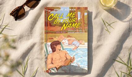 „Call me by your name“ liegt mittig auf einem hellen Tuch. Oben links liegt eine Sonnenbrillen, oben rechts steht ein Glas mit Zitronenscheibe. Gras und Schatten umgeben das Buch.