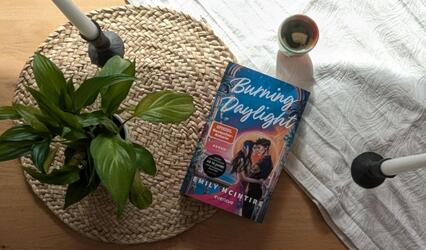 „Burning Daylight“ liegt auf einem Tisch. Darunter liegen ein Tischset aus Bast und ein weißer Tischläufer. Daneben stehen eine kleine Topfpflanze sowie zwei Stabkerzen.