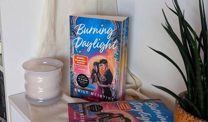 Auf einem weißen Regal steht das Buch „Burning Daylight“ aufrecht. Links daneben steht eine helle Kerze. Unten liegt ein zweites Buch. Rechts ragt eine grüne Pflanze ins Bild. Der Hintergrund ist ein heller Stoff.