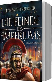 Das Buchcover zeigt einen kriegerischen römischen Soldaten mit einer markanten roten Helmzier. Der Hintergrund ist in dunklen Farben gehalten und zeigt schemenhafte Figuren von weiteren Soldaten, die eine dramatische Atmosphäre erzeugen. Der Titel „Die Feinde des Imperiums“ ist in großen, goldenen Buchstaben prominent platziert, während der Untertitel „Historischer Roman“ in kleinerer, silberner Schrift darunter steht. Der Name des Autors, Jens Wittenberger, befindet sich oben auf dem Cover.