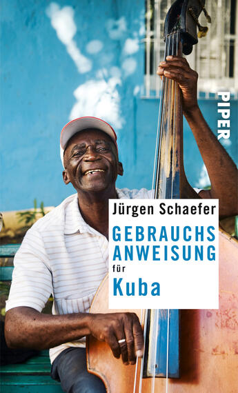Das Buchcover zeigt einen lachenden älteren Mann, der einen Kontrabass spielt. Er trägt ein weißes Hemd und eine graue Mütze. Im Hintergrund ist eine blauen Wand sowie eine Holzbank zu sehen, die eine entspannte Atmosphäre vermittelt. Der Titel „GEBRAUCHSANWEISUNG für Kuba“ steht in großen, weißen Buchstaben auf einem blau-weißen Hintergrund, gefolgt vom Autorennamen Jürgen Schaefer. Das Verlagslogo „PIPER“ befindet sich oben rechts.