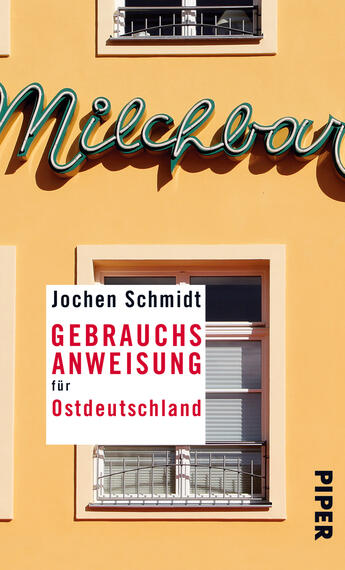 Das Buchcover zeigt eine gelbe Wand mit einem großen, nostalgischen Schriftzug „Milchbar“ in grünlicher Farbe. Darunter ist ein Fenster mit einem Gitter und vor einem weißen Hintergrund ein rotes Rechteck, auf dem der Titel „GEBRAUCHSANWEISUNG für Ostdeutschland“ steht. Der Autor „Jochen Schmidt“ ist in Schwarz oberhalb des Titels platziert, und das Verlagssignet „PIPER“ befindet sich in der unteren rechten Ecke. Das Design vermittelt eine warmherzige, kulturelle Atmosphäre.