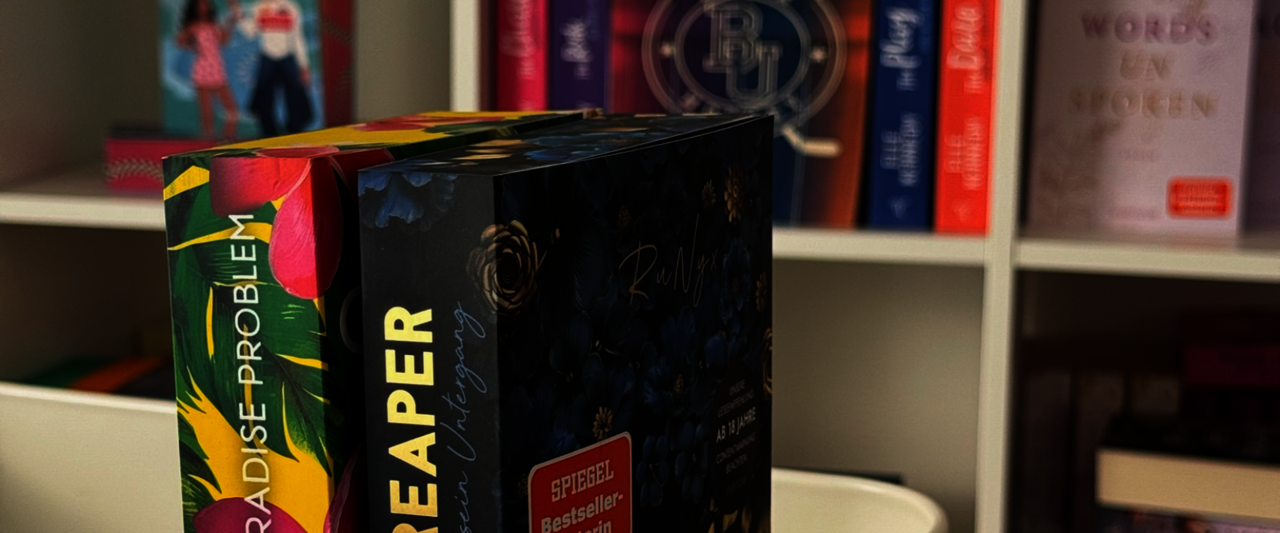 Im Vordergrund stehen zwei Bücher: „The Reaper“ mit einem floralen, dunklen Cover und „The Paradise Problem“ mit buntem, tropischem Design. Im Hintergrund befinden sich Bücher in einem Regal.