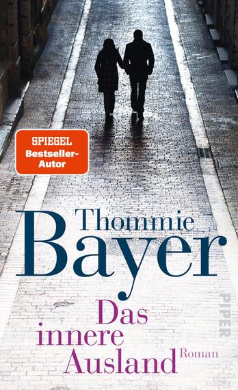 Das Buchcover zeigt zwei silhouettenhafte Figuren, die Hand in Hand auf einem schmalen, gepflasterten Weg gehen. Der Hintergrund ist in sanften Grautönen gehalten und vermittelt eine nostalgische Atmosphäre. Oben links befindet sich ein rotes Feld mit dem Text „SPIEGEL-Bestseller-Autor“. Der Titel „Das innere Ausland“ ist in großer, blauer Schriftzentriert, während der Autorennamen „Thommie Bayer“ in eleganten, größeren Buchstaben darunter steht. Der Schriftzug wird durch den Verlagsnamen ergänzt.