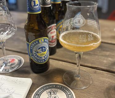 Ein Stielglas mit Berliner Weisse steht auf einem Holztisch. Im Hintergrund sind fünf verschiedene Bierflaschen der Brauerei Lemke Berlin sichtbar und im Vordergrund ein Notizbuch sowie ein Bierdeckel mit der Aufschrift „Geschmack ist unser Bier“.