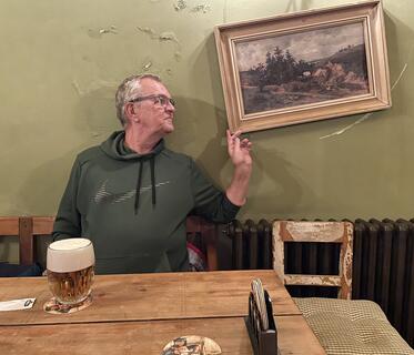 Ein Mann in einem dunkelgrünen Pullover sitzt vor einer hellgrünen Wand an einem Holztisch. Vor ihm steht ein voller Bierkrug aus Glas, und er zeigt auf ein schräg hängendes Landschaftsgemälde neben ihm an der Wand.