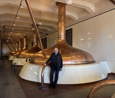 Fünf raumhohe Kupferkessel stehen hintereinander in einer traditionellen Brauerei. Sie sind auf weiß gefliesten Podesten platziert. Das helle Interieur hat eine Kachelwand und eine Decke mit kleinen Lampen. Davor steht ein Mann in dunkler Kleidung und mit Brille.