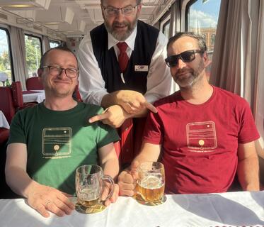 Zwei Männer sitzen an einem Tisch in einem Zugrestaurant. Vor ihnen stehen kleine Bierkrüge aus Glas. Ein Kellner steht hinter ihnen und zeigt mit einer freundschaftlichen Geste auf die beiden Männer. 
