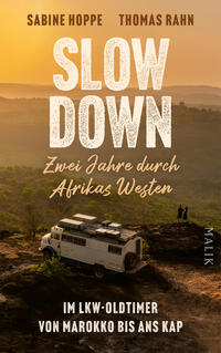 Das Buchcover zeigt eine Szene im goldenen Abendlicht: oben die Autorennamen Sabine Hoppe und Thomas Rahn in blockiger, braun‑goldener Versaltypografie. Zentral prangt der Titel SLOW DOWN in großen, rauen weißen Großbuchstaben, darunter die kursiv-handschriftliche Zeile „Zwei Jahre durch Afrikas Westen“. Im Vordergrund ein weißer LKW-Oldtimer auf einem Felsen, zwei winzige Silhouetten, weiter Blick über nebelige Hügel, ruhige, abenteuerliche Atmosphäre.