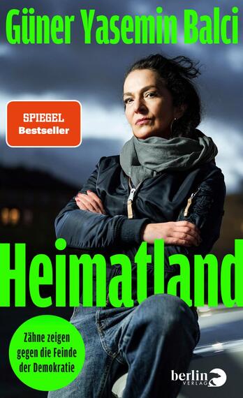Das Buchcover zeigt eine Porträtaufnahme einer Frau, die ernst in die Kamera blickt. Sie trägt einen schwarzen Winterjacke und eine graue Schal. Der Hintergrund ist in düsteren, graublauen Tönen gehalten, was eine nachdenkliche Stimmung erzeugt. Der Titel „Heimatland“ ist in großen, leuchtend grünen Buchstaben platziert. Darunter steht der Untertitel „Zähne zeigen gegen die Feinde der Demokratie“ in einer runden, grünen Blase. Der Autorinnenname „Güner Yasemin Balci“ ist oben auf dem Cover in schlichtem Weiß abgedruckt.