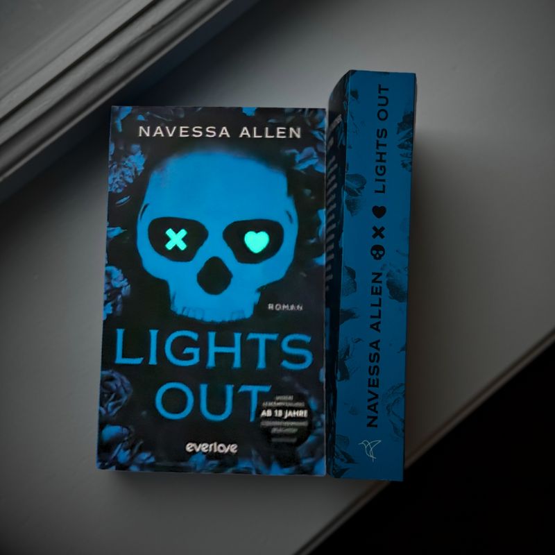 "Lights Out" von Navessa Allen liegt einmal mit dem Cover nach oben und einmal mit dem Buchrücken nach oben auf einem weißen Fensterbrett. Der Leuchteffekt auf dem Cover ist zu sehen.