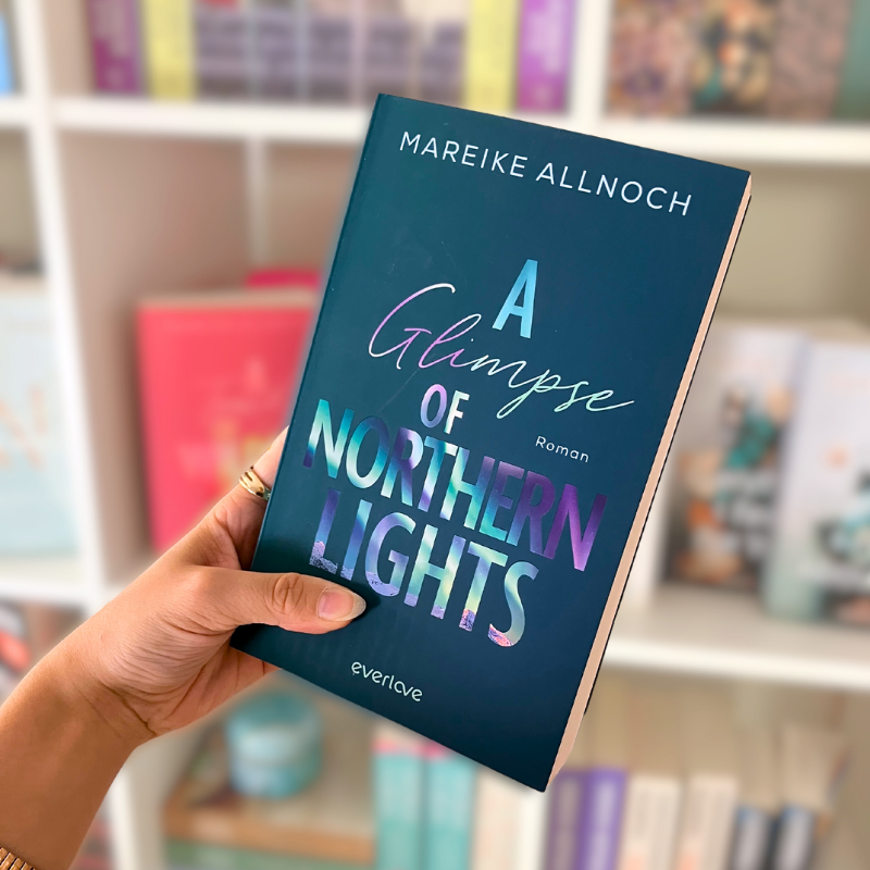 „A Glimpse of Northern Lights“ von Mareike Allnoch wird vor einem Bücherregal gehalten