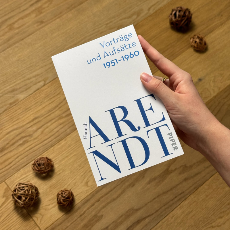 „Vorträge und Aufsätze 1951–1960“ von Hannah Arendt in einer Hand über einem Holzboden gehalten.