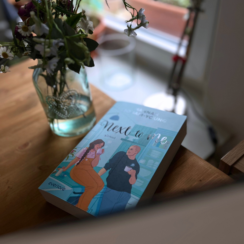 „Next to Me“ liegt auf einem Holztisch neben einer Vase mit Blumen