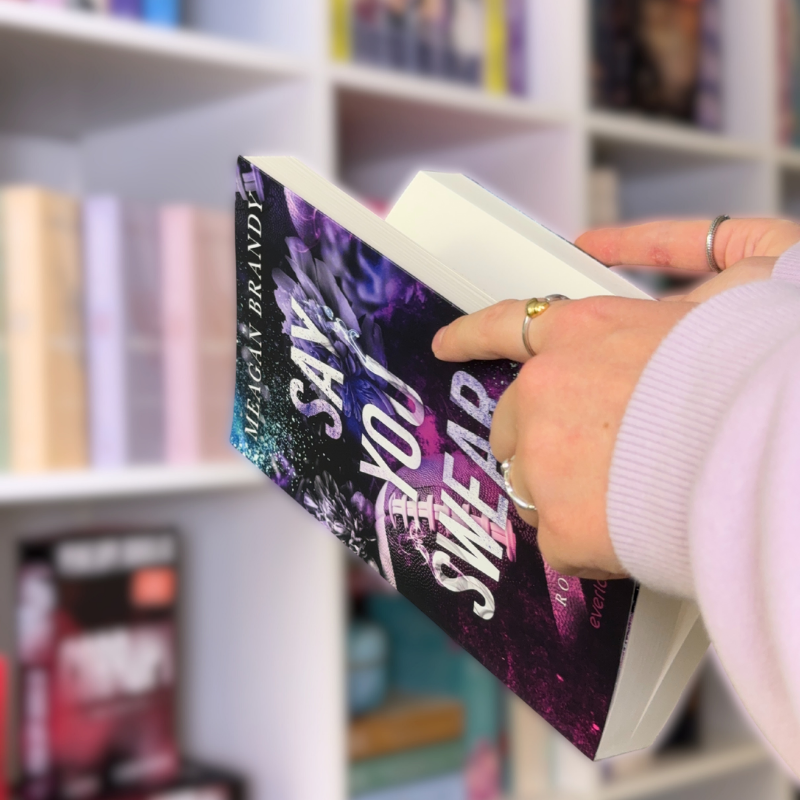 „Say You Swear“ von Meagan Brandy wird durchgeblättert. Im Hintergurnd steht ein Bücherregal