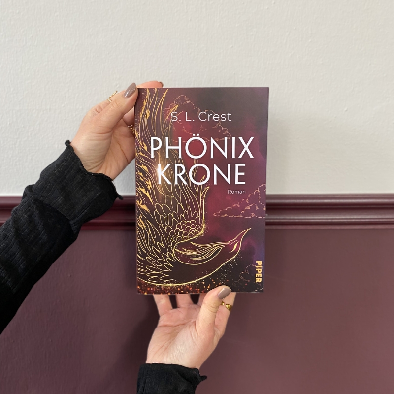 "Phonixkröne" wird von zwei Händen vor einer Wand gehalten, deren obere Hälfte weiß und deren untere Hälfte weinrot ist