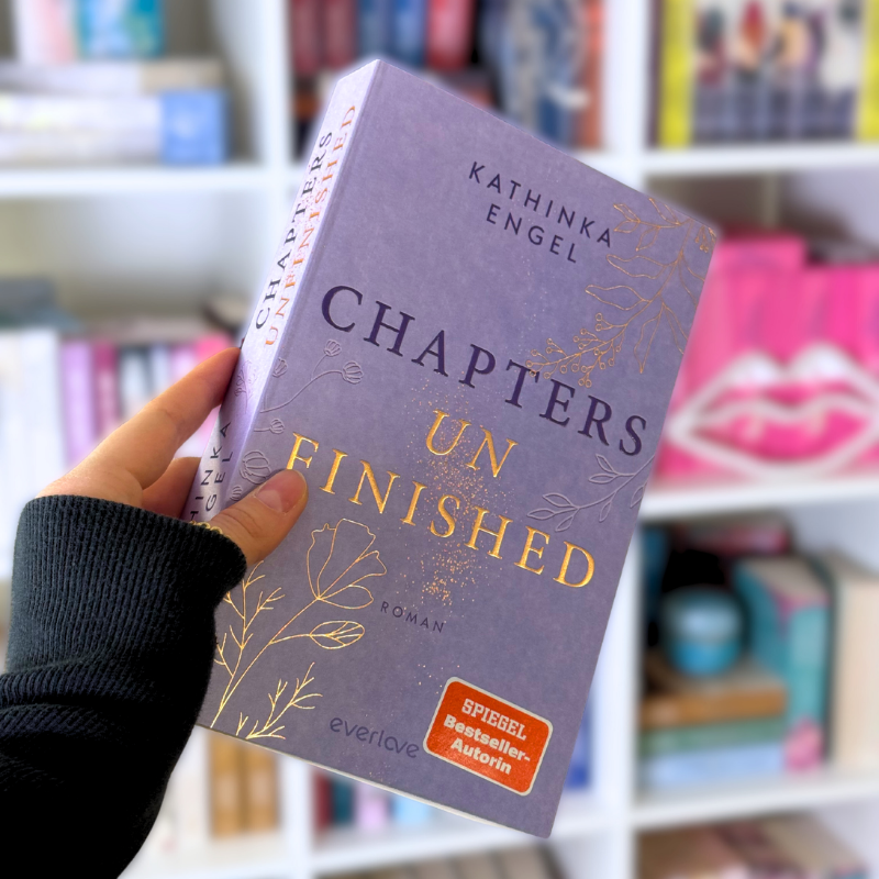 „Chapters Unfinished“ von Kathinka Engel wird vor einem Bücherregal gehalten.