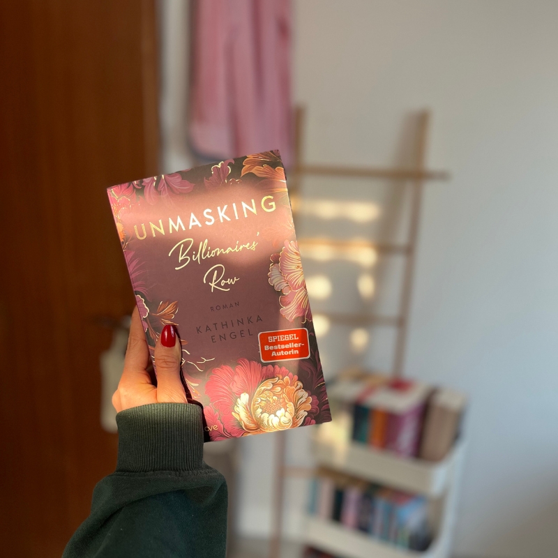 Eine Hand hält "Unmasking Billionaires' Row". Die Sonne scheint darauf und im Hintergrund ist ein kleiner Bücherwagen zu sehen.