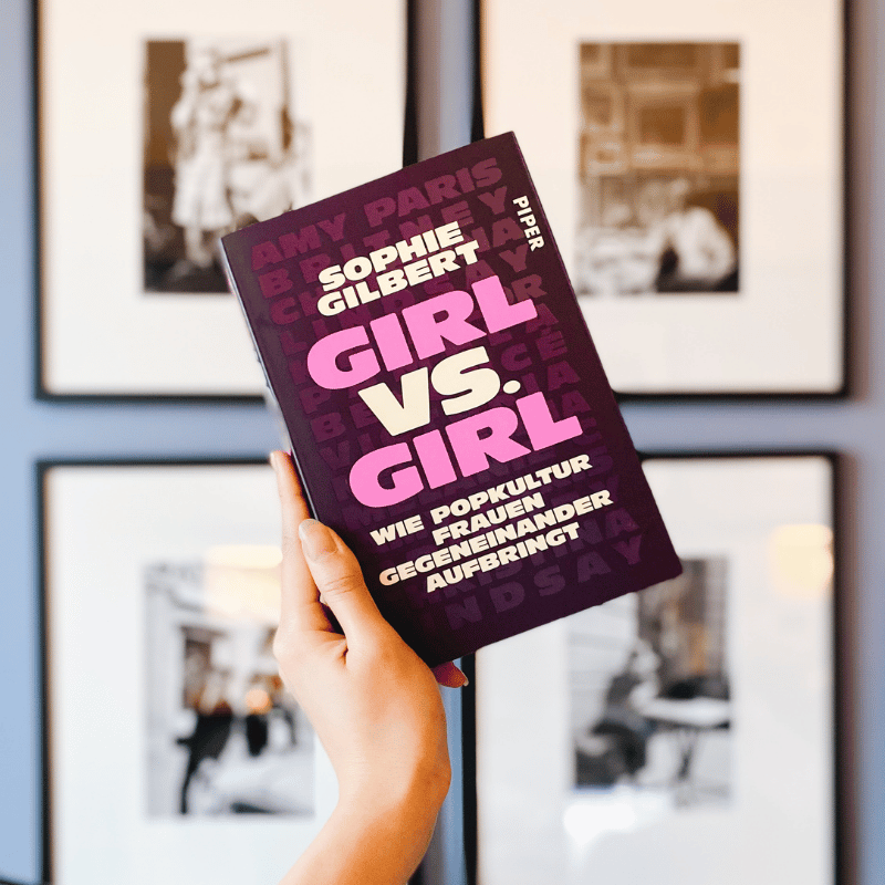Eine Hand hält das Buch „Girl vs. Girl“ von Sophie Gilbert vor einer Wand mit Bildern hoch